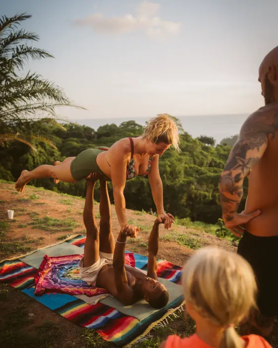 A woman balancing flat on a partner's feet in acro yoga on the land at Amora, the forest canopy and the Pacific behind.