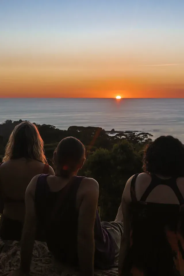 Three people sit together on a blanket on a grassy rise at Amora, looking out over treetops to the sun setting into the Pacific — the ocean-view ridge of the ecovillage above Dominicalito, Costa Rica.