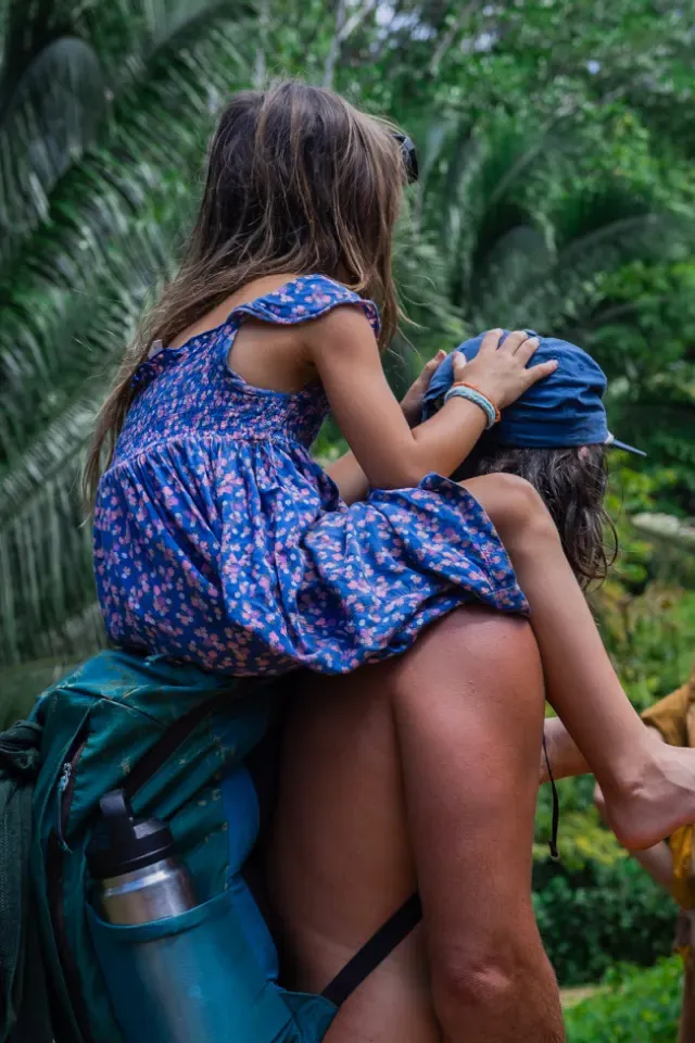 A child in a blue floral dress rides on an adult's shoulders through dense palm forest on the land at Amora, near Dominical, Costa Rica.