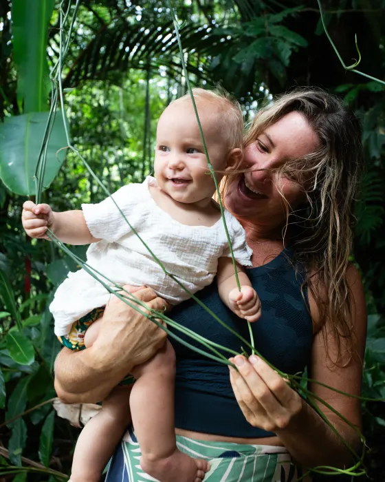 A laughing woman holding a baby among wide jungle leaves and hanging vines on the land at Amora.